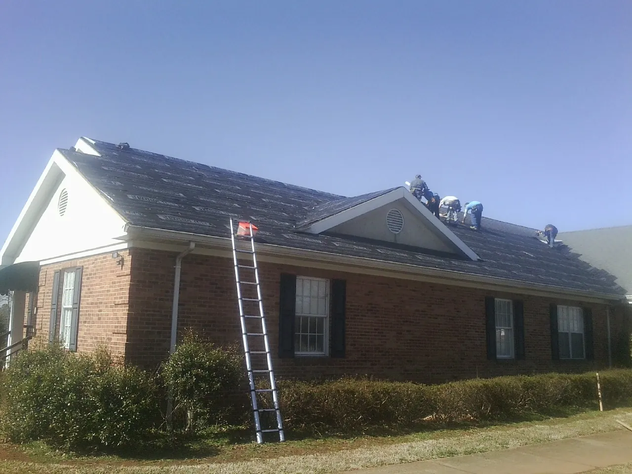 Crew tearing shingles off a brick ranch roof with a ladder set — Emergency Roof Repair work by our crew in West Grove