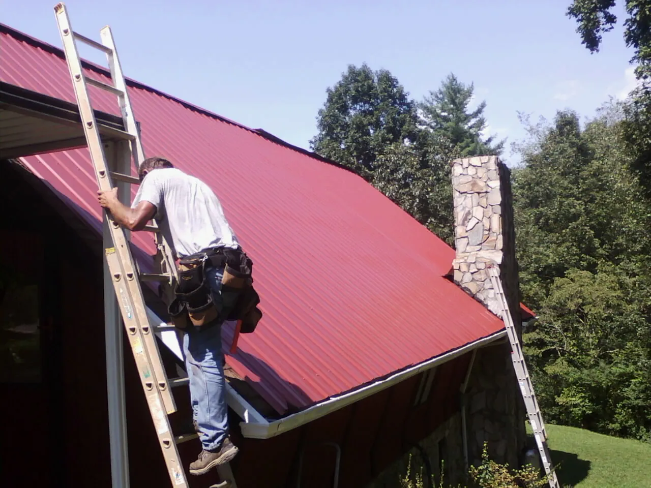Roofer on a ladder beside a red metal roof and stone chimney — Soffit & Fascia Repair by Roofing Brothers in West Grove